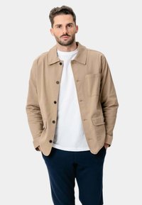 Beige jakke lavet af bomuld med krave, knaplukning og en brystlomme. Båret over en hvid skjorte med navy bukser.