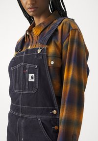 Mørke denim overaller med justerbare stropper, med en frontlomme og messingdetaljer, kombinert med en rutete, flerfarget flanellskjorte.