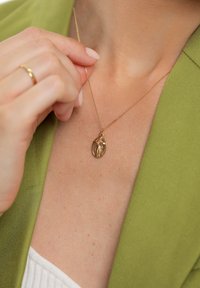 Guldfärgat halsband med korshänge, fäst vid en fin kedja; bärs över en ljus topp mot en grön kavaj.