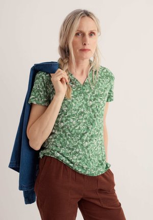 Frau mit blondem Zopf trägt ein grün gemustertes Hemd und braune Hose, hält eine blaue Jacke über der Schulter und schaut nach vorne.