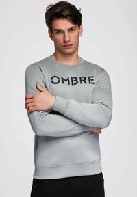 Młody mężczyzna o ciemnych włosach, ubrany w jasno szarą bluzę z napisem "OMBRE", stojący ze skrzyżowanymi ramionami, patrzący prosto w obiektyw na jednolitym tle.