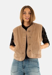Gilet di velluto a coste beige con colletto alla mandarino, chiusura con cinque bottoni e tasche laterali. Indossato sopra un top nero a maniche lunghe.