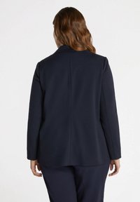 Blazer navy cu mâneci lungi și rever tăiat, având un material neted și o croială ajustată, prezentând un design simplu pe spate, cu detalii minime.