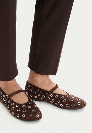 Brązowe zamszowe baleriny Mary Jane z srebrnymi oczkami, noszone z ciemnobrązowymi, krótkimi spodniami.