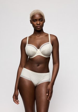 Vrouwelijk model met kort blond haar draagt een bijpassende crèmekleurige beha en hooggetailleerde slip met subtiele geometrische patronen, poserend tegen een effen achtergrond.