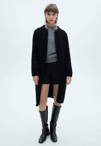 Cappotto lungo nero e soffice sopra un maglione grigio, abbinato a pantaloncini neri e stivali neri al ginocchio. Tessuto liscio e vestibilità comoda.