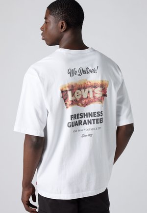 Mężczyzna zwrócony bokiem, ubrany w białą koszulkę z grafiką kawałka pizzy i tekstem „Dostarczamy! Gwarancja świeżości Levi's.”