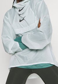 Chaqueta Nike gris claro con un alto cuello y tres logotipos negros en el frente. Presenta mangas largas y sueltas y un diseño en capas sobre una camisa de color teal.