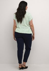 Top verde a maniche corte con un motivo astratto bianco, abbinato a pantaloni fitted blu scuro, visto da dietro. Scarpe nere flat completano il look.