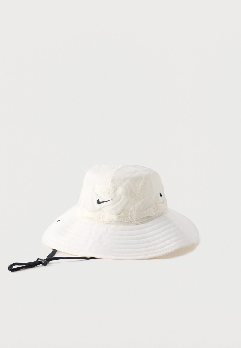 Λευκό καπέλο του τύπου bucket με φαρδιά γείσα, με μαύρο λογότυπο Nike Swoosh και ρυθμιζόμενο μαύρο λουράκι κάτω από το σαγόνι σε απλό φόντο.