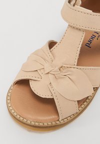 Sandale en cuir beige avec un détail en nœud, bout ouvert et sangle en velcro. Elle possède une semelle en caoutchouc de couleur naturelle et des accents cousus.