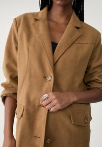 Blazer marron texturé avec une fermeture à un bouton, des revers crantés et des poches latérales. Associé à une bague spirale argentée portée à la main.