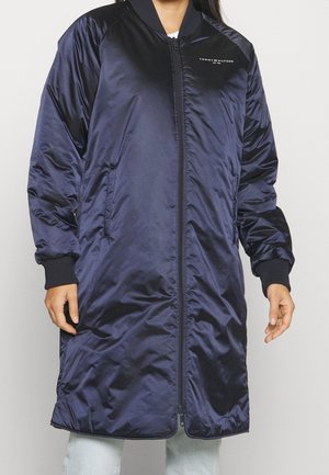 Blouson aviateur long en satin bleu marine avec fermeture éclair à l'avant, poignets côtelés, poches latérales et petit logo Tommy Hilfiger sur la poitrine porté par une personne.