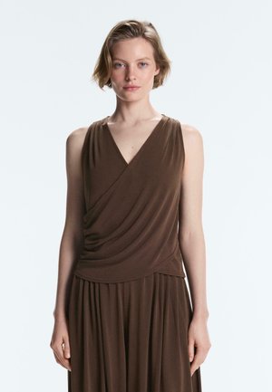 Rövid szőke hajú fiatal nő barna, ujjatlan, átkötős stílusú felsőt és hozzáillő pliszírozott szoknyát visel, fehér háttér előtt áll.