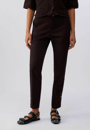 Person iført mørke, tætsiddende bukser, en mørk tekstureret top og sorte flade sko med nitter og dobbelt rem, der står mod en ensfarvet baggrund.