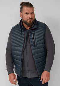 Dunkelblaues Pufferwesten mit horizontalem Steppmuster, vorderem Reißverschluss und kleiner Reißverschlusstasche. Über einem grauen Zip-Hoodie getragen.
