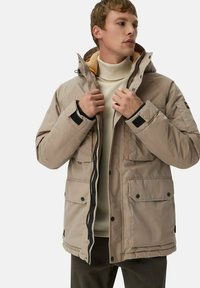 Beige isolierte Jacke mit Kapuze, Frontreißverschluss und Druckknopftaschen. Verfügt über ein kontrastierendes orangenes Futter und schwarze Akzente an den Ärmeln.