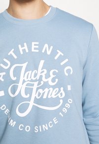 Světlomodrá mikina z bavlněného materiálu s velkým bílým nápisem „Jack & Jones“ a kruhovými akcenty.