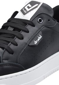 Sneaker in pelle nera con suola in gomma bianca, lacci neri piatti e dettagli perforati. Presenta un logo bianco prominente sulla linguetta.