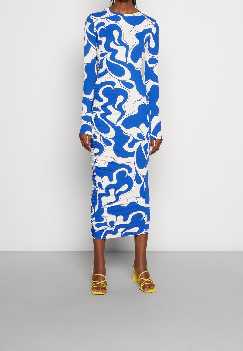 Robe à manches longues présentant un audacieux motif abstrait bleu et blanc, avec des détails de fronces sur les côtés et une silhouette ajustée. Associée à des sandales jaunes.