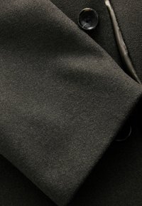 Mörk ullkappa i tyg med en mjuk textur, med släta ärmdetaljer och en glänsande svart knappaccent.