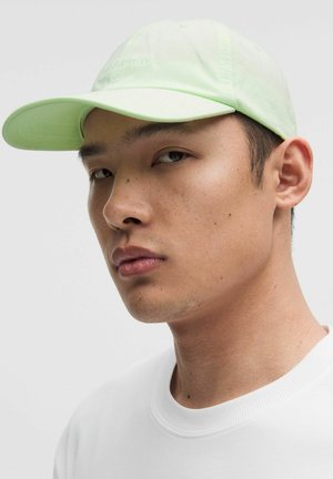 Jonge man met een lichtgroene baseballpet en een witte shirt met ronde hals, kijkt iets naar de zijkant tegen een effen achtergrond.