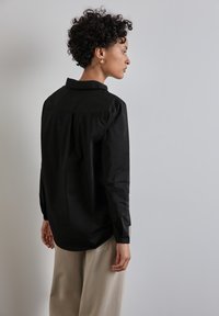 Camicia nera con collo a camicia, maniche lunghe e texture liscia, caratterizzata da una vestibilità comoda e un orlo curvo. Indossata con pantaloni beige.