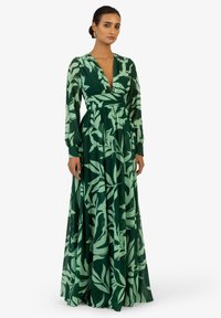 Robe longue verte avec motif de feuilles vert clair. Présente un décolleté en V profond, des manches longues et une taille cintrée, s'épanouissant en une jupe ample. Tissu lisse.