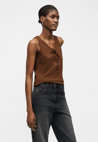Γυναίκα που φοράει αμάνικη καφέ μπλούζα με λεπτομέρειες δέσιμου μπροστά και σκούρο τζιν με ψηλή μέση, στέκεται μπροστά σε απλό φόντο.