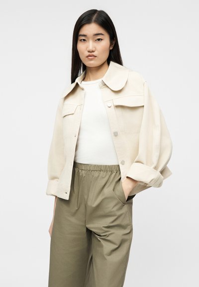 Mlada ženska z dolgimi temnimi lasmi, oblečena v kremno jakno preko belega vrhnjega dela in olivno zelenih hlač z elastičnim pasom, z eno roko v žepu.