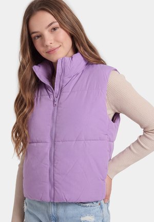 Jonge vrouw met lang haar die een lichtpaarse gewatteerde bodywarmer draagt over een beige geribd shirt met lange mouwen en lichtblauwe spijkerbroek.