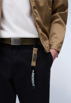 Personne portant une veste beige, une chemise blanche, un pantalon noir avec une ceinture de marque et un porte-clés accroché à la boucle de ceinture du côté droit.