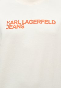 Λευκό ύφασμα με έντονη πορτοκαλί επιγραφή "KARL LAGERFELD JEANS" τυπωμένη στην περιοχή του στήθους.