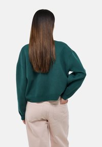 Pull vert oversize avec une coupe décontractée, des poignets côtelés et un ourlet détaillé, associé à un pantalon beige clair. Texture lisse, design décontracté.
