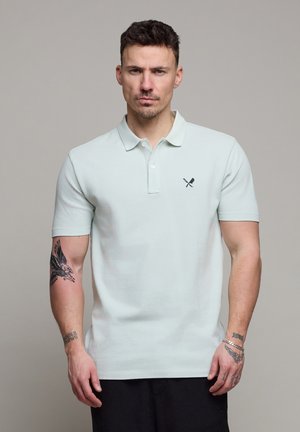 Man met kort donker haar, gekleed in een lichtblauw poloshirt met een logo van een gekruiste mes en spatel, met tatoeages op beide armen zichtbaar.