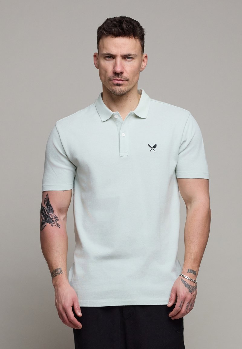 Homme aux cheveux courts et foncés portant un polo bleu clair avec un logo de couteau croisé et spatule, montrant des tatouages sur les deux bras.