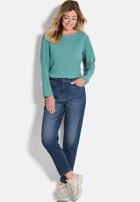 Haut turquoise à manches longues côtelé avec un col rond, associé à un jean bleu taille haute et des baskets beiges. Texture douce, coupe décontractée.