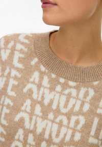 Personne portant un pull en maille beige avec des lettres abstraites blanches, montrant le cou et le bas du visage légèrement tournés sur le côté.