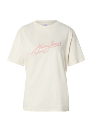 Femme blonde aux yeux bleus portant un t-shirt oversize crème avec l'inscription rose « Nani Jeans » et un jean bleu clair sur fond gris.