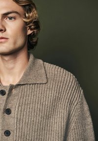 Cardigan en tricot côtelé beige avec un col, doté de boutons foncés et d'un tissu texturé, présenté sur un fond neutre.