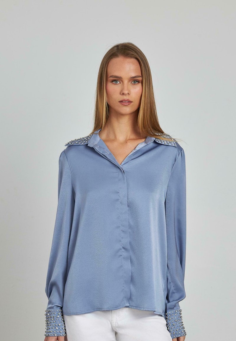 Blusa di raso azzurro chiaro con maniche lunghe, decorata con borchie argentate sulle spalle e sui polsini, con chiusura a bottoni davanti e colletto.