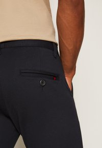 Gros plan d'une personne portant un pantalon noir avec une poche arrière boutonnée et un haut beige, debout devant un fond uni.