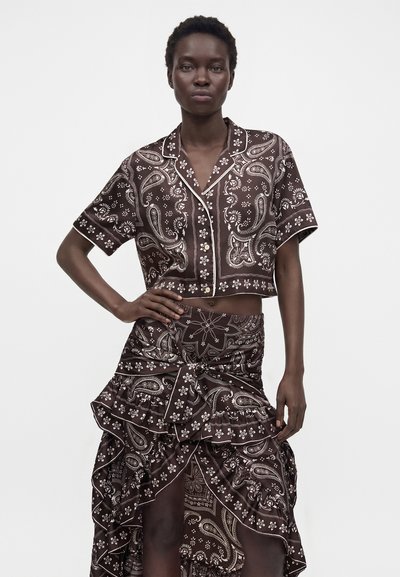 Femme portant une chemise boutonnée à manches courtes et imprimé paisley foncé assorti, et une jupe asymétrique à volants, main sur la hanche, sur fond blanc.