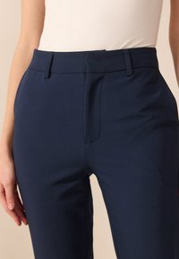 Námornícky modré nohavice so strihom na mieru a hladkým povrchom, s prednými skladmi, pútkami na opasok a zipsovým zapínaním. Materiál je mierne elastický.