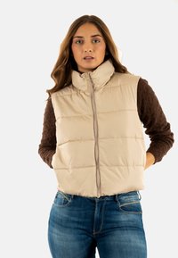 Chaleco acolchado beige con cuello alto, frente con cremallera y patrón acolchado. Combinado con una blusa de manga larga marrón y texturizada y unos vaqueros.