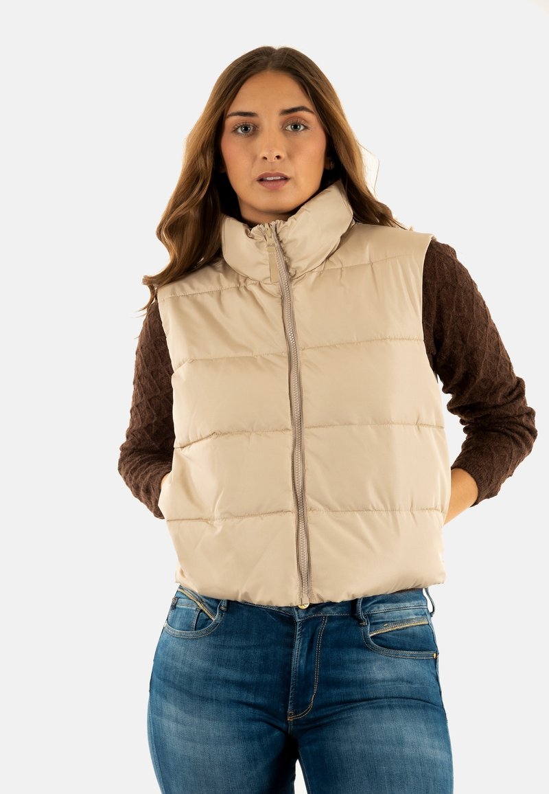 Chaleco acolchado beige con cuello alto, frente con cremallera y patrón acolchado. Combinado con una blusa de manga larga marrón y texturizada y unos vaqueros.