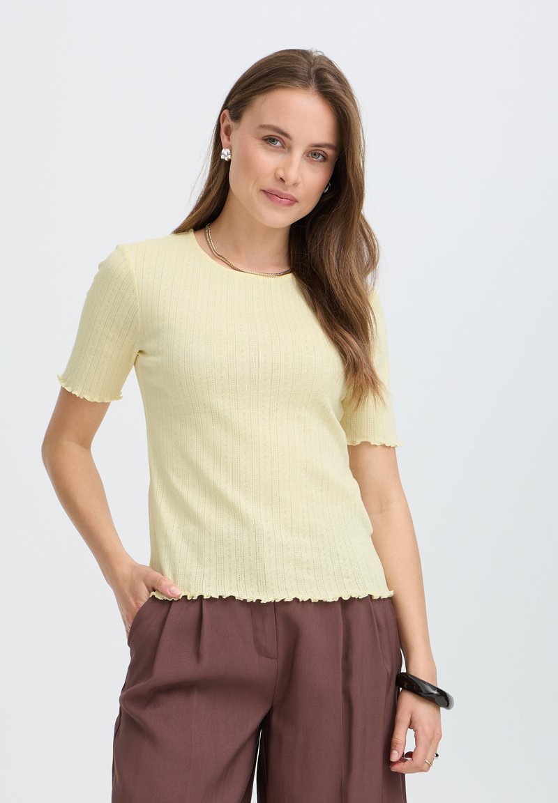 Femme aux longs cheveux bruns portant un haut côtelé jaune pâle à manches courtes et un pantalon marron, debout avec une main dans la poche.