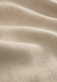 Svijetlo bež laneni materijal s mekanom, glatkom teksturom, koji se odlikuje suptilnim nerazmjernostima u tkanju i varijacijama boje.