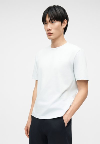 Jeune homme aux cheveux noirs portant un t-shirt blanc uni à manches courtes et un pantalon noir, debout avec une main dans la poche sur fond blanc.