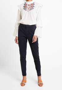 Vit blus med volangärmar och broderade detaljer i röd och blått, tillsammans med marinblå byxor och orange klacksandaler.